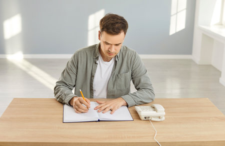Young man being busy while searching job, making calls via stationary phone and writing down information in notepad, sitting at desk in empty room, having serious expression, trying to find workの写真素材