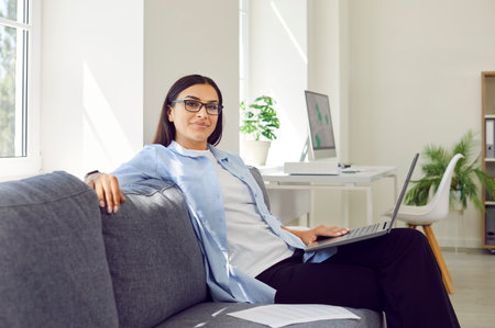 Portrait of young businesswoman sitting on sofa and working on laptop. Beautiful relaxed lady in casual clothes and eyeglasses sitting on couch and looking at cameraの写真素材