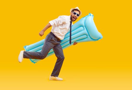 Happy, joyful, funny, excited man office worker in white shirt, trousers, tie, sun hat and sunglasses holding blue rubber beach mattress and running on yellow studio background. Summer holiday conceptの写真素材