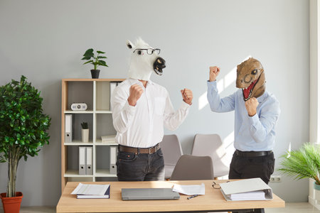 Two businessmen in dinosaur and horse animal masks raising their hands up in office. Two business partners, colleagues celebrating their success, complete project, got great newsの写真素材
