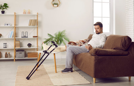 Man with plastered leg sitting on couch using mobile phone at home. Young injured man with fractured leg in cast staying at home. Rehabilitation and orthopaedic recovery conceptの写真素材