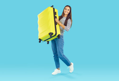 Cheerful positive tourist woman carrying yellow heavy suitcase. Happy girl wearing striped long sleeve and jeans going to travel for summer vacation. Traveling and tourism conceptの写真素材