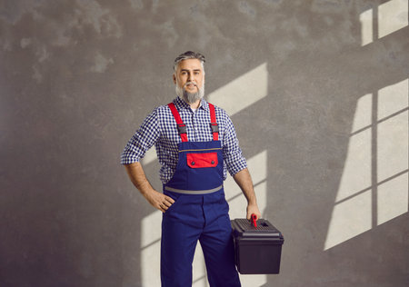 Portrait of a mature smiling bearded repairman holding a tool box in his hands on a gray wall background. Professional gray-haired handyman worker standing cheerful in blue overall.の写真素材