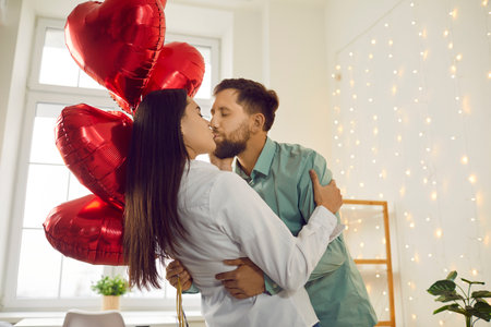 Young happy smiling brunette man holding red foil ballons in his hand and kissing pretty woman on holiday and hugging. Happy couple in love. Care, tenderness and Valentines day concept.の写真素材