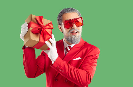 Merry Christmas and Happy New Year concept. Thoughtful grey haired Santa Claus wearing bright red costume, sunglasses and white gloves, holding gift box, isolated over vivid green backgroundの写真素材