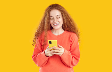 Smiling redhead curly girl in red sweatshirt looking at smartphone screen on yellow background. She swipes and scrolls on screen of mobile phone. Chatting, online, working or socializing remotely.の写真素材