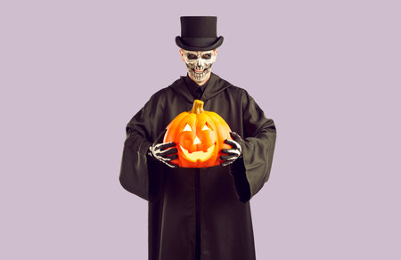 Portrait of man with face painted for Halloween holding pumpkin. Spooky man wearing black cloak, top hat and gloves standing on isolated studio background. Day of the Dead, Halloween conceptの写真素材