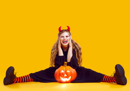 Young girl in carnival costume of devil with horns sitting in the studio on an orange background with a Halloween pumpkin and screaming with open mouth and closed eyes with shocked face expression.の写真素材