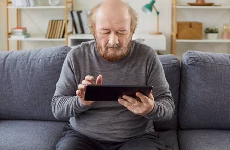 Bearded senior man trying to understand how to use modern technologies, holding digital tablet, surfing internet, reading news online, posing on comfortable sofa in cozy living room.の写真素材