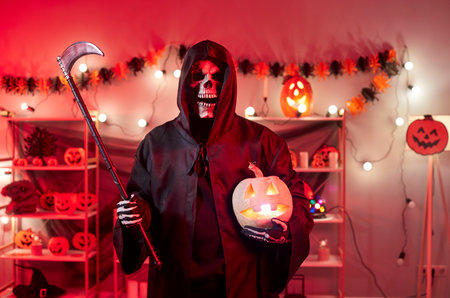 Portrait of male person in grim reaper death costume in black cloak with hood looking at camera holding scythe and pumpkin in living room at home. Halloween party celebration and horror concept.の写真素材