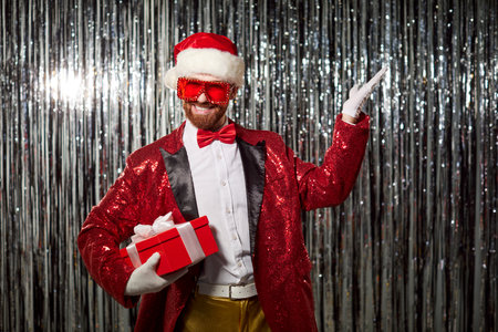 Portrait of positive man in red Santa costume holding gift box, enjoying fun during Christmas or new year evening party in a festive setting with warmth and celebration every moment.の写真素材