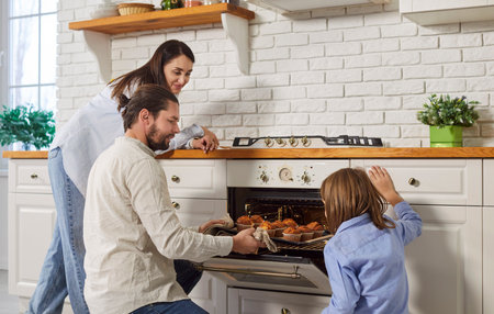 Happy family, young couple and kid taking tray with yummy self baked tasty muffins out of oven, enjoy cooking in modern kitchen, homemade family pastries, culinary hobby, cupcakes dessert, tasty hotの写真素材