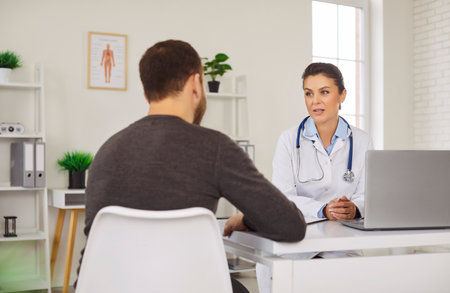 Health care. Female doctor conducts one-on-one consultation with patient in modern medical office. Young doctor wearing white coat listens attentively to concerns of patient sitting in front of desk.の写真素材