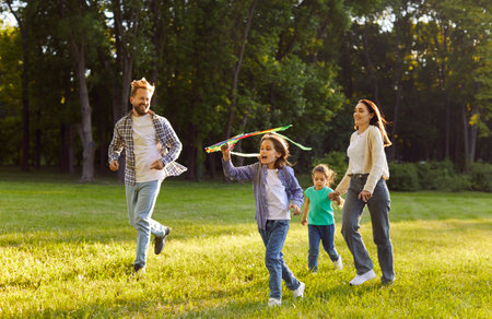 Outdoor shot of happy friendly family flying kite in park during windy day, spending free time outside. Father and mother playing with their children, having entertainment in summerの写真素材