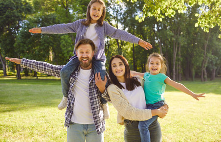Cheerful father giving piggy back to his son, lovely mother carrying daughter on hands while spending free time in park. Happy family enjoying nice spring day outdoors. People and leisureの写真素材