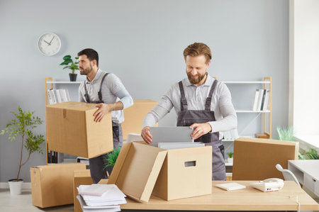Moving delivery pack service, male movers team moving cardboard boxes, loading, unloading setting up of household goods, new house stuff, carrying stacks of cardboard relocation boxes in office roomの写真素材