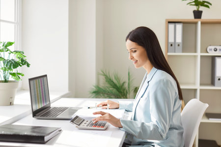 Woman working online in office using laptop and calculator for business accounting and documents audit, focusing on precision in a professional setting with attention to detail and analysis.の写真素材