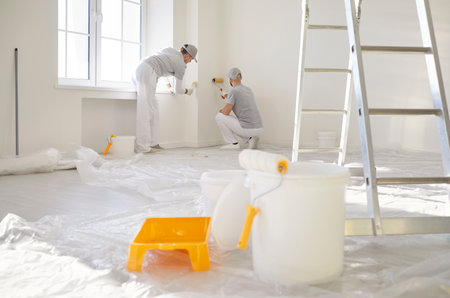 Paint roller, buckets, tray and ladder symbolizing renovation process in progress with two painters working in background. Workers applying fresh paint to walls with rollers during construction works.の写真素材