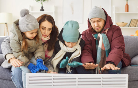 Family freezing in extreme cold, warming near heater at home, suffering in cool flat, shivering tremble with cold, unhappy people wearing upper clothes feel badly, bad room heating system problemsの写真素材