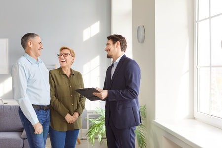 Happy smiling mature senior couple discussing with young man realtor real estate contract going to sign document. Elderly man and woman having consultation with financial advisor standing indoors.の写真素材