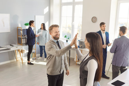 Man company employee giving high five with woman celebrating success, making a deal, business achievement or signing a contract on workplace. Group of business people working in officeの写真素材