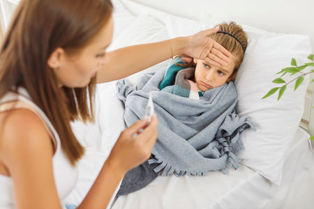 Caring kind young affectionate mom and sick child in bed measuring temperature, touching forehead,...
