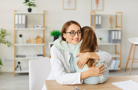Portrait of a friendly smiling pediatrician woman hugging with a little cute child girl sitting in medical clinic after examination. Female doctor with kid patient in office. Health care concept.の写真素材