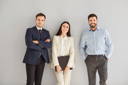 Portrait of a group of three young cheerful business people in suits standing confidently, looking at the camera and smiling isolated on a studio grey background. Team work concept.の写真素材