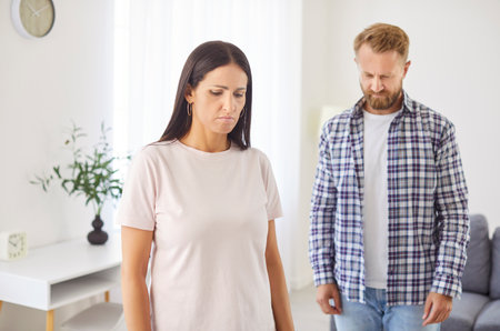 Upset young family couple standing in silence in living room after quarrel. Emotionally drained husband and wife showing frustration and sadness after arguing, reflecting deep relationship problems.の写真素材
