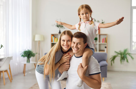 Happy playful loving family portrait, smiling laughing young parents with cute little daughter together posing in modern living room at home, relaxing on weekend to rest, care and love, having funの写真素材