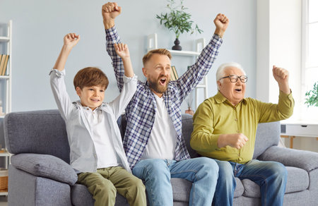 Three generations of men cheer energetically watching exciting sports competitions on TV. Little boy cheering with his father and grandfather sitting on sofa at home and raising hands in celebration.の写真素材