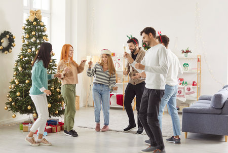 Group of young friends dancing in living room with decorated Christmas tree holding glasses of champagne. Happy people celebrating New Year party at home. Celebration and winter holidays concept.の写真素材