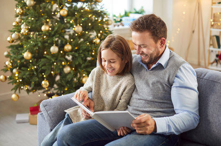 Happy smiling child looking through a family photo album sitting on sofa at home with young father enjoying Christmas or New Year eve together. Family leisure and winter holiday concept.の写真素材