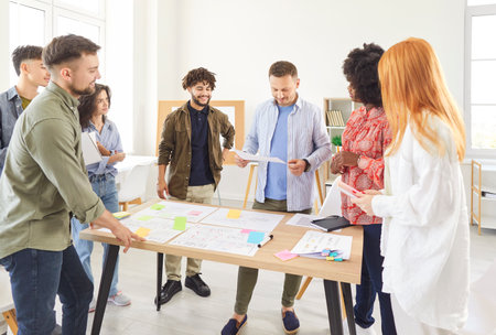 Multiracial group of young people in casual wear working together in collaborative project. Diverse team of students or colleagues standing around desk, brainstorming, discussing ideas, planning work.の写真素材