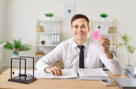 Portrait of a happy businessman working in a modern office, holding a light bulb sticker to represent a new idea. Moment of inspiration, eureka and creativity in a professional setting.の写真素材