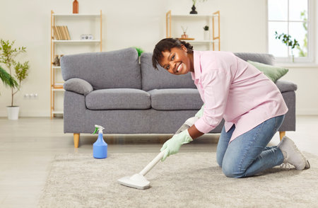Portrait of a happy african american woman vacuuming the carpet on floor while cleaning cozy and comfortable house. Maid in household chores, maintaining a tidy and organized home.の写真素材