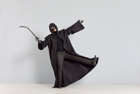 Unknown person in Halloween costume representing Death poses spectacularly with scythe in right hand. Full length of man wearing black robe with hood and skull mask against white wall background.の写真素材