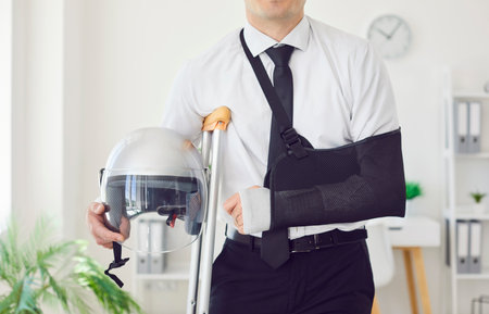 Midsection shot of man stands in office, with broken, bandaged arm supported by crutch, suffering by injury. Holding helmet, he exemplifies resilience in face of trauma and accidents.の写真素材