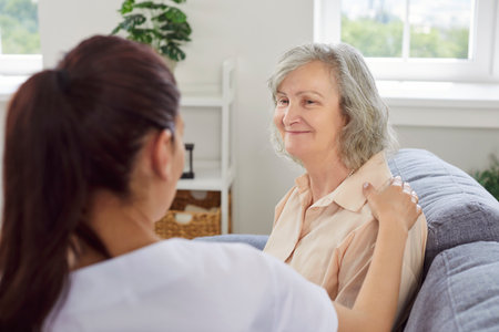 Senior woman patient sitting on sofa and talking with female young friendly nurse visiting her at home during medical examination. Doctor support and nursing for retirement people concept.の写真素材