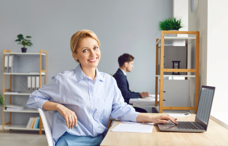 Portrait of a young confident business woman sitting at the desk on workplace, looking at the camera and smiling. Cheerful accountant working on a laptop with tables and charts at office.の写真素材