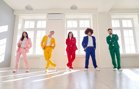 Full length portrait of confident young people in bright colorful suits standing in a row indoors and looking at camera. Cheerful guys and girls in fashionable stylish party outfit.の写真素材