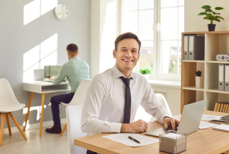 Portrait of young happy smiling attractive business man typing on a laptop at office and looking cheerful at camera. Male cheerful company employee working at the desk on his workplace.の写真素材