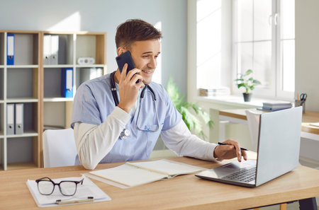 Portrait of a young male doctor in blue medical uniform sitting at the desk using laptop working in medical office. Man physician talking by mobile phone on his workplace in clinic.の写真素材