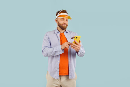 Connected and casual. Portrait of cheerful focused man in casual clothes engrossed in his smartphone. Smiling millennial man in sporting visor scrolls mobile phone screen on light blue background.の写真素材