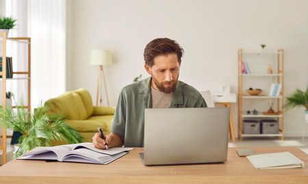 Portrait of confident bearded young business man in casual clothes working at the desk on his workplace with laptop at office or at home making notes in his notepad looking at computer monitor screenの写真素材
