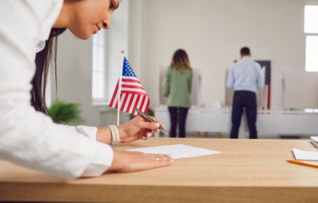 Young woman employee work in voting center. People american voters voting standing back in a row at vote center with USA flags in polling booth. Election day and democracy concept.の写真素材