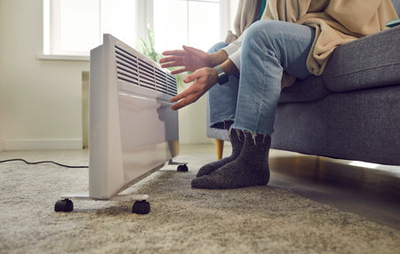 Cropped photo of a frozen male person sitting on the sofa in the living room wrapped in blanket at home and trying to warm his hands near modern electric heater. Heating problems concept.の写真素材