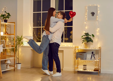 Valentines day. Young happy woman holding red foil balloon in hand hugging and congratulating boyfriend on holiday. Smiling couple man and woman in love standing at home looking at each other.の写真素材