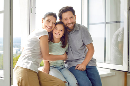 Portrait of smiling young happy family of three with daughter sitting on the windowsill at home and looking cheerful at camera hugging their child girl. Love, care, family time and leisure concept.の写真素材