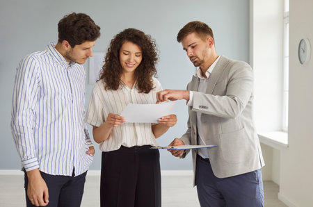 Portrait of confident man professional realtor standing with happy couple clients in modern new apartment discussing contract buying property. Move, rent and real estate market concept.の写真素材